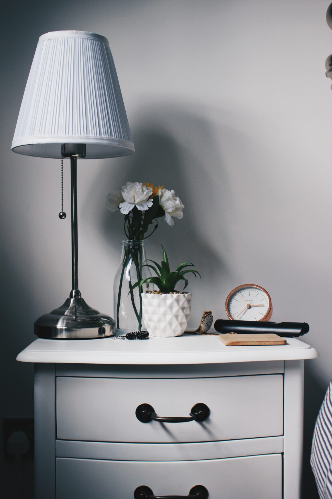 White bedside table decorated with a lamp, flowers, alarm clock, and remote