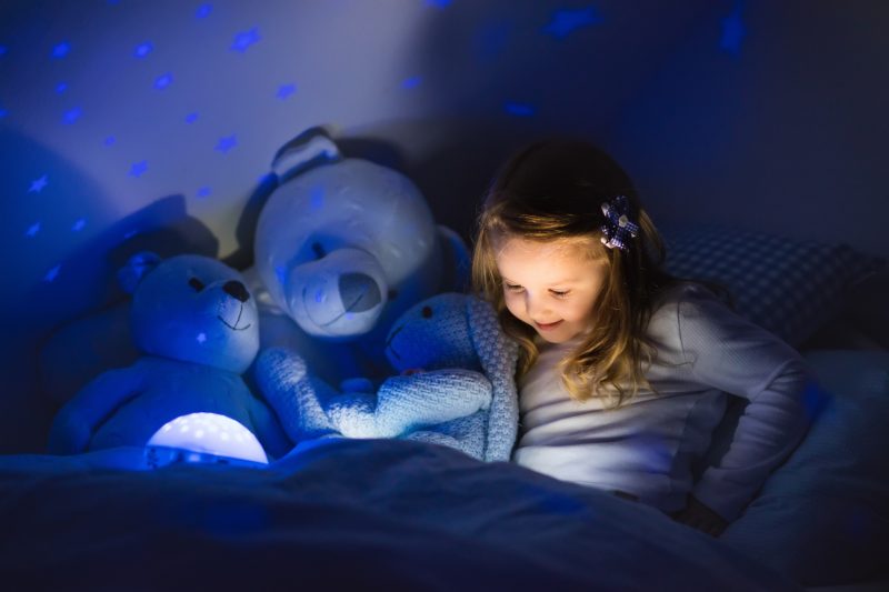 Little girl reading a book with her stuffed animals in a dark bedroom lit by a night light projecting stars onto the wall