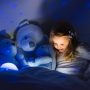 Little girl reading a book with her stuffed animals in a dark bedroom lit by a night light projecting stars onto the wall