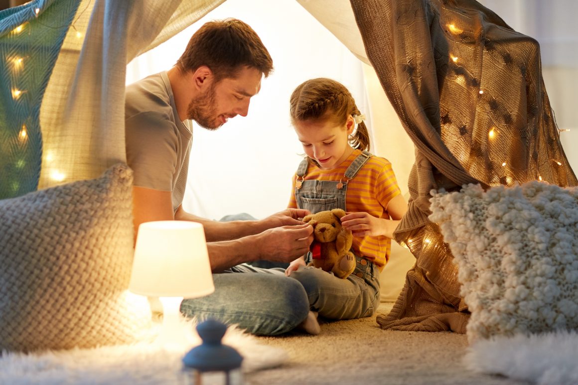 Dad playing in a warmly lit homemade blanket and pillow fort with his young daughter